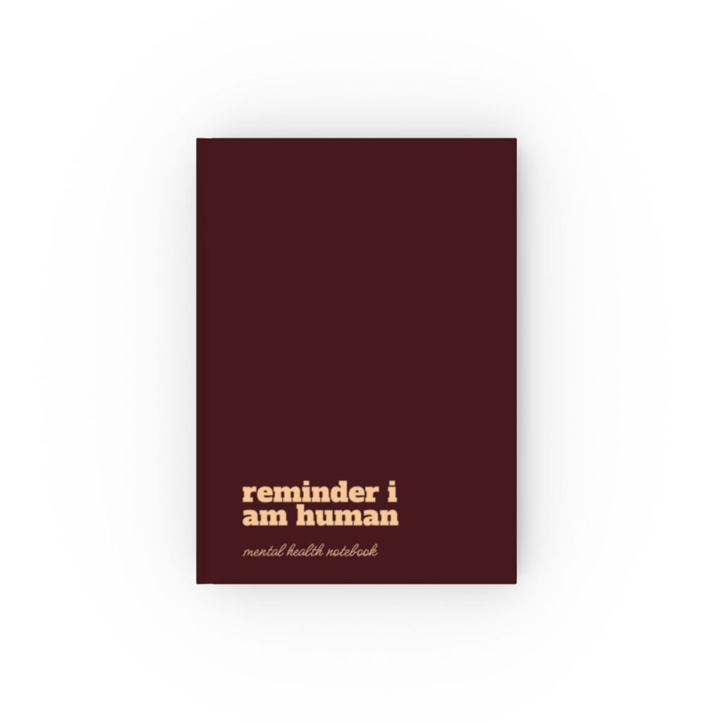 An deep maroon hardcover notebook cover with bold peach text reading "reminder i am human." Below, smaller text says "mental health notebook." The tone is empowering by iziiexpressions.com