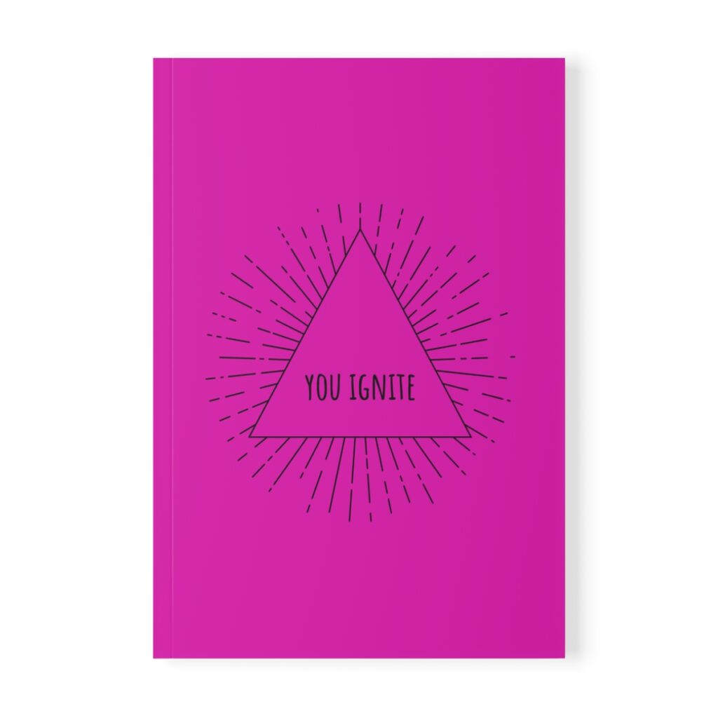 Pink softcover notebook with a black triangle in the center. Inside the triangle, the text reads "You Ignite" with radiating lines around it, conveying motivation by iziiexpressions.com