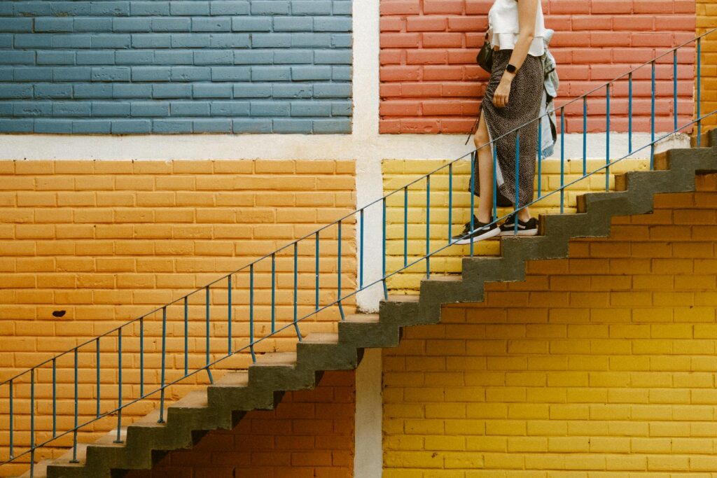 A person descends stairs against a vibrant wall divided into blue, red, yellow, and orange sections, creating a lively, geometric backdrop. Photo by Diego Marcel: https://www.pexels.com/photo/woman-walking-downstairs-near-colorful-wall-25682344/