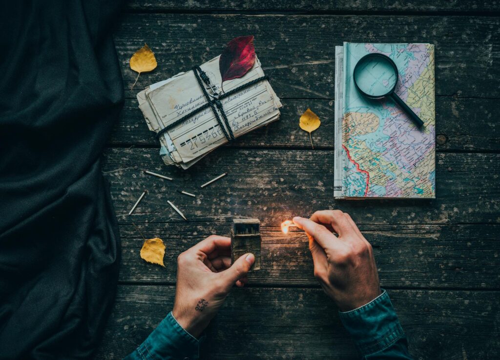 Hands lighting a match next to scattered letters tied with string and a leaf, surrounded by a map with a magnifying glass, on a rustic wooden table signifies travel experiences. Photo by Elijah O'Donnell: https://www.pexels.com/photo/person-holding-a-lighted-matchstick-3088778/
