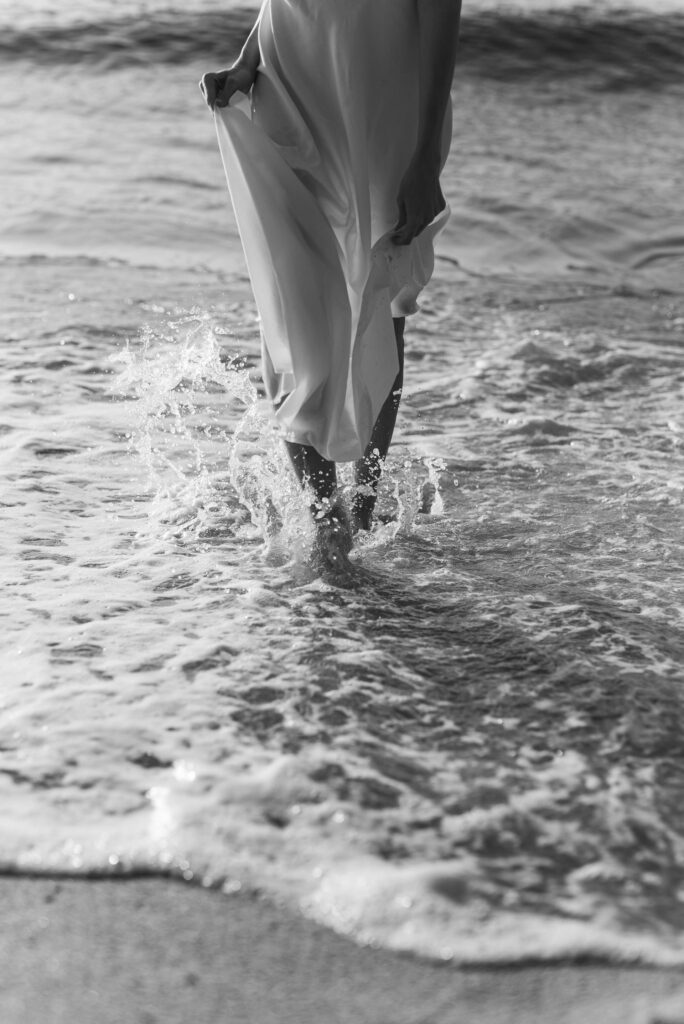 A person in a flowing dress walks along the shoreline, feet splashing through gentle waves. The black-and-white image conveys a serene, contemplative mood. Photo by Leeloo The First: https://www.pexels.com/photo/a-person-walking-on-the-shore-8908810/