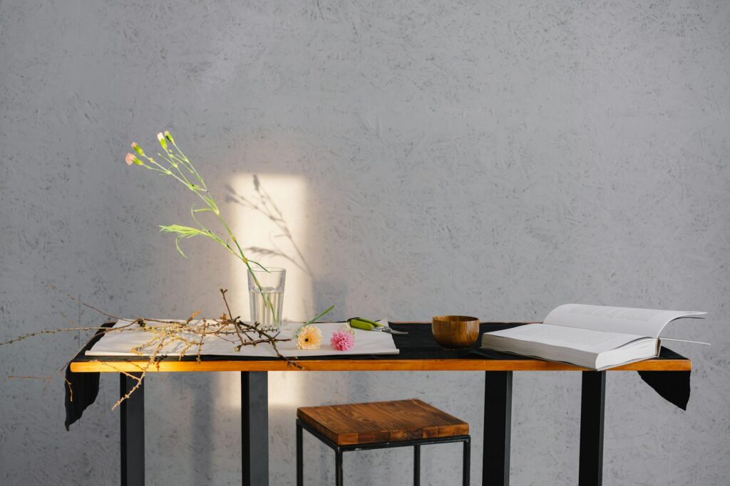 Minimalist table scene with open book, glass with flowers, wooden bowl, and branch casting shadow on textured gray wall. Calm, serene atmosphere. Photo by Mikhail Nilov: https://www.pexels.com/photo/brown-wooden-table-with-white-printer-paper-on-top-6933429/